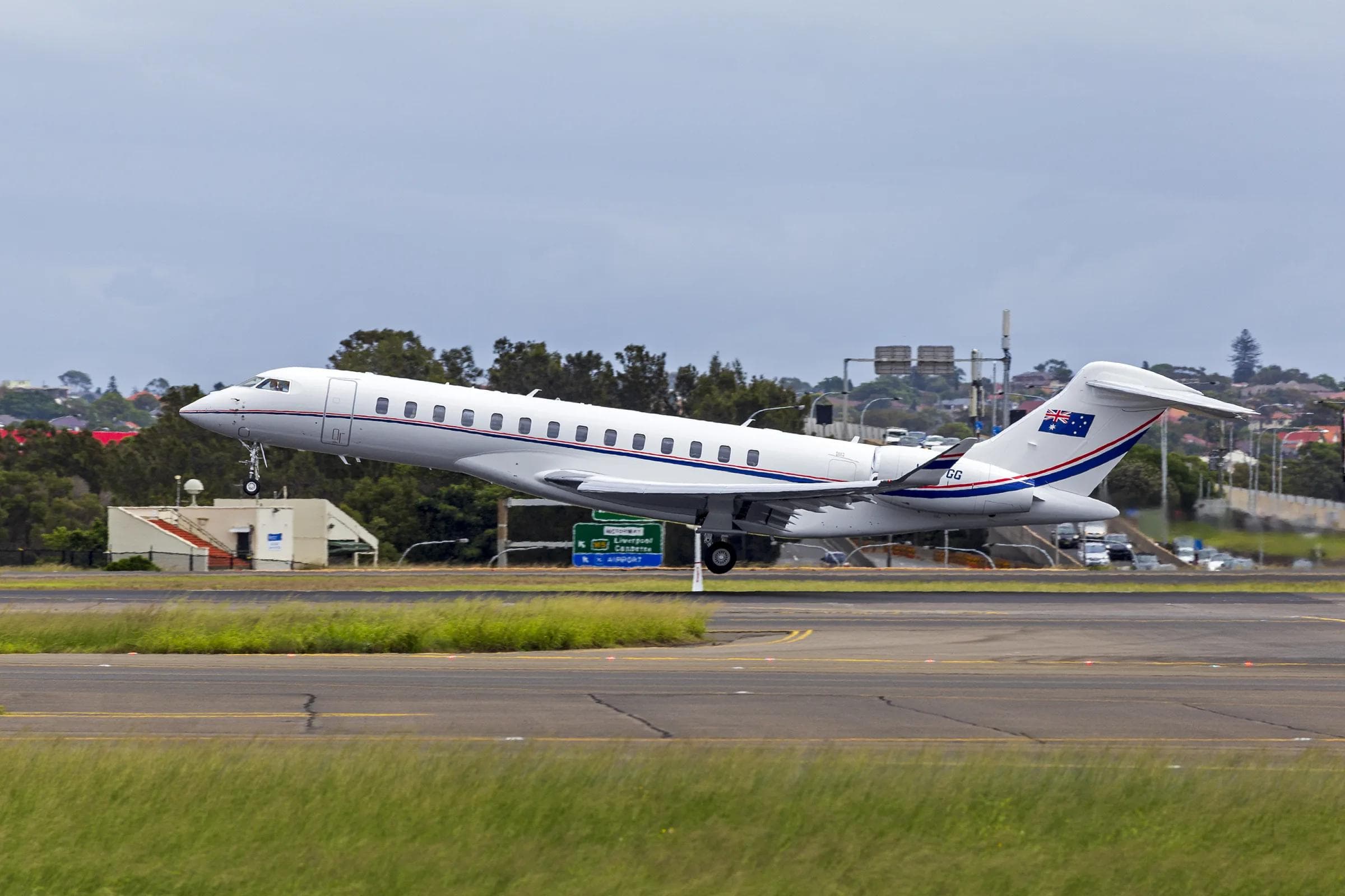 Bombardier Global 7500 business jet in takeoff rotation at Sydney Airport, white livery with Australian flag tail marking, grey sky
